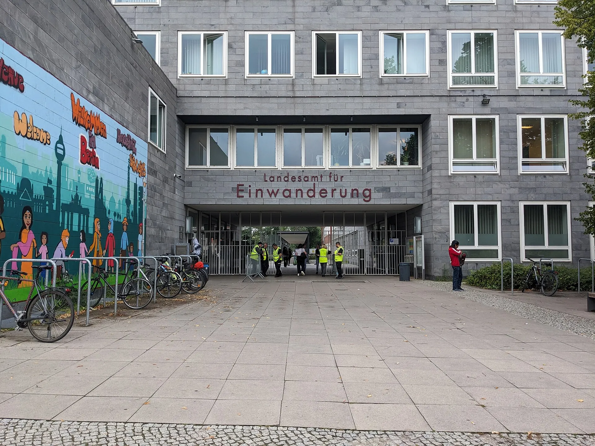 Ingresso del Landesamt für Einwanderung di Berlino, con la scritta sulla facciata, biciclette parcheggiate e un murale di benvenuto in più lingue