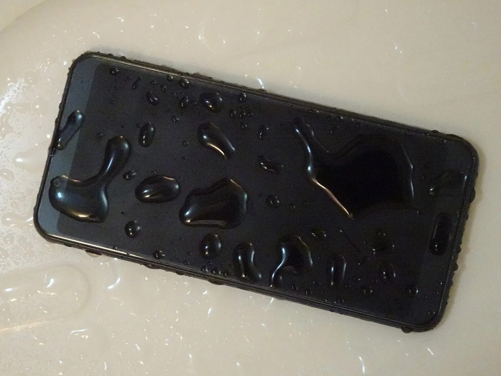 Smartphone nero coperto di gocce d’acqua, appoggiato sul fondo di un lavandino bianco