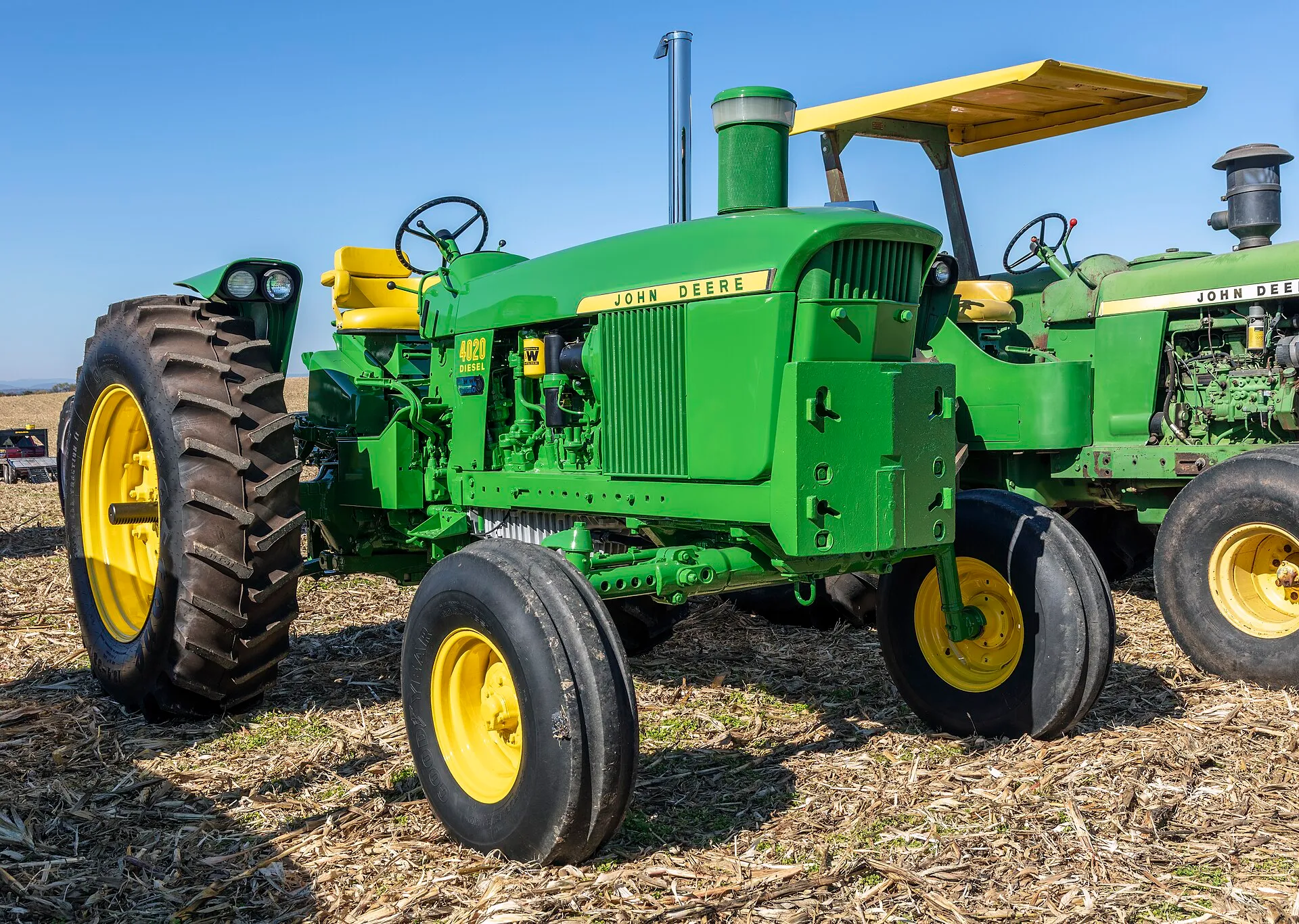 Trattore John Deere 4020 Diesel verde con cerchi gialli, parcheggiato su un campo di stoppie accanto ad altri trattori d’epoca